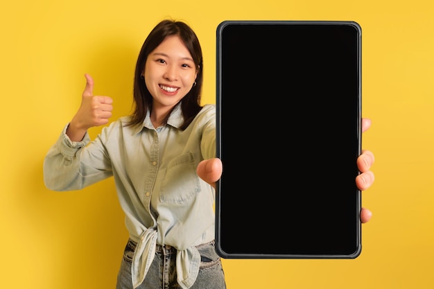 Young asian woman showing tablet with empty screen gesturing thumb up offering mockup for website or