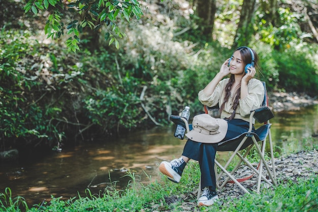 Молодая азиатка сидит в кресле возле ручья, слушает музыку в беспроводных наушниках и с удовольствием использует планшет во время кемпинга в лесу