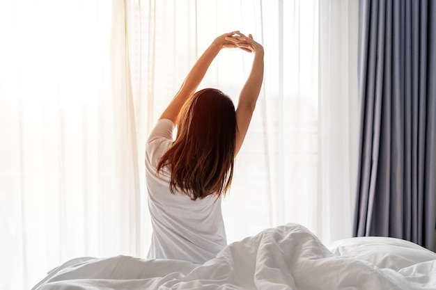 Молодая азиатская женщина сидит на кровати и растягивается утром