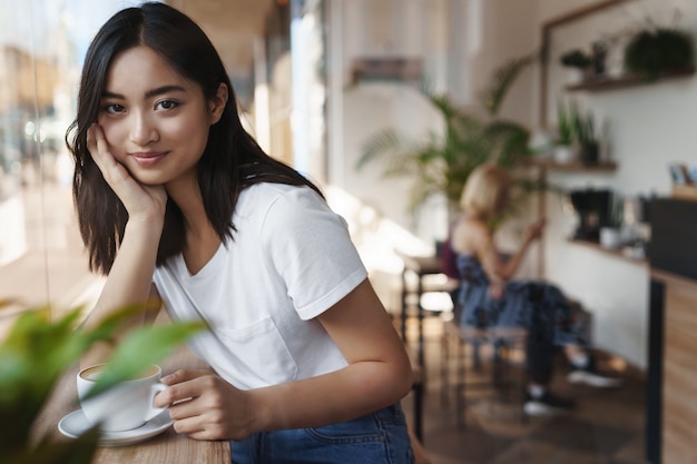 窓の近くのカフェに座って、コーヒーを飲む若いアジア人女性。