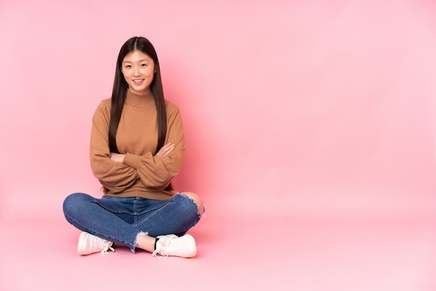 Young asian woman sitting on the floor on pink wall keeping the arms crossed in frontal position