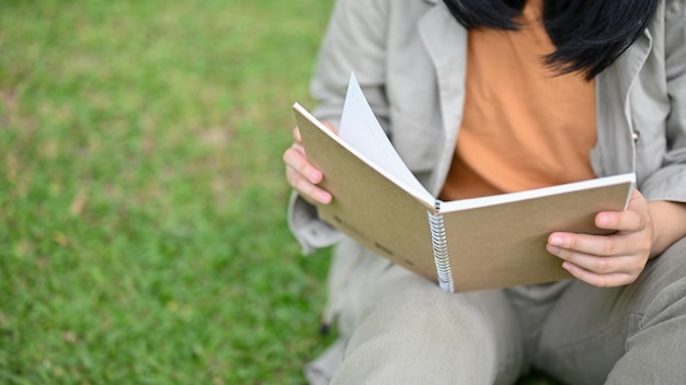 Giovane donna asiatica che si siede sull'erba verde nel parco che si rilassa e che legge un libro