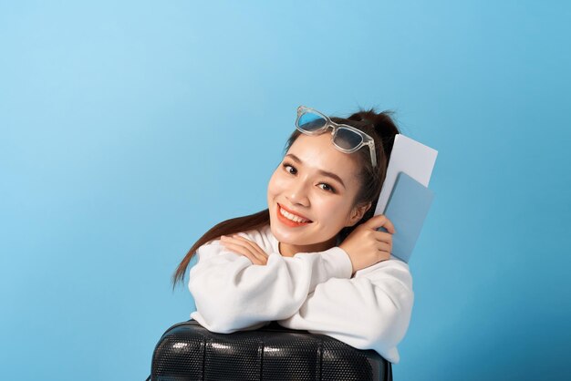 Young asian woman sitting near a suitcase holding passport on a blue background