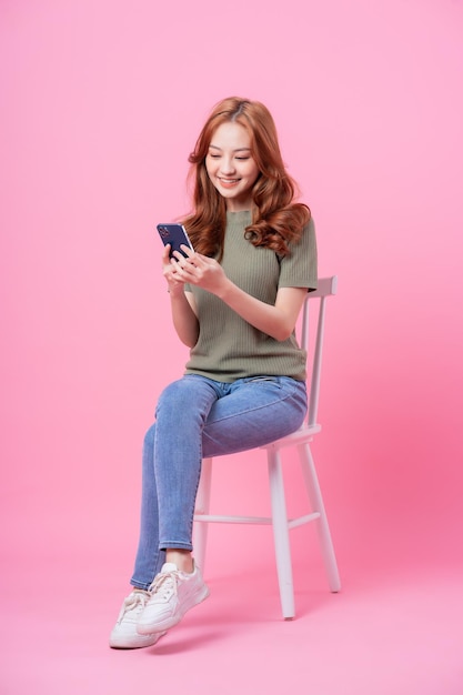 Young Asian woman sitting and using smartphone on pink background