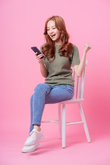 Young Asian woman sitting and using smartphone on pink background