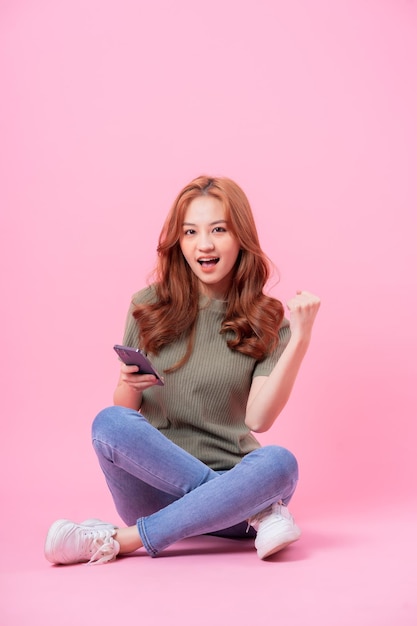 Young Asian woman sitting and using smartphone on pink background