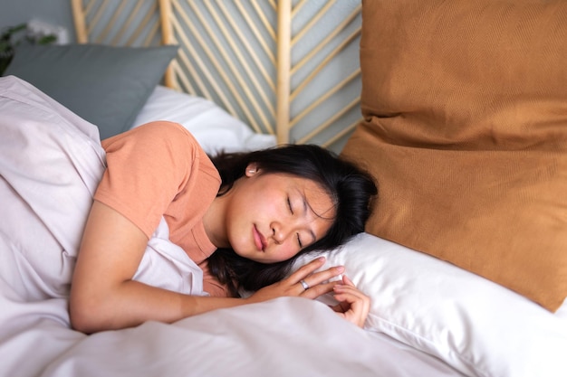 Молодая азиатка спит в уютной постели с большим количеством подушек и удобным одеялом.