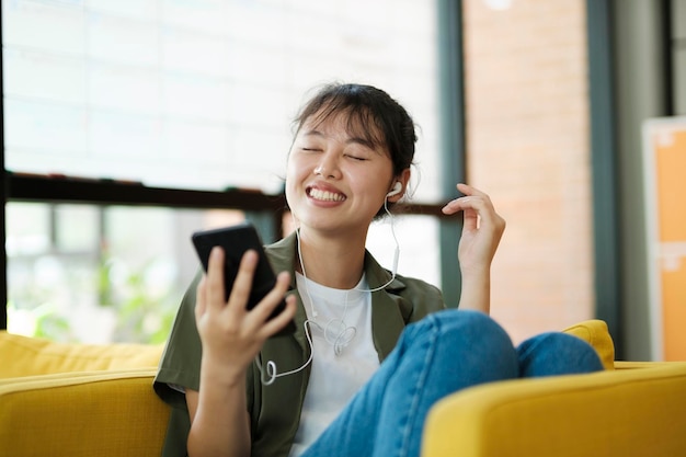 Giovane donna asiatica sorridente ascoltando musica utilizzando lo smartphone a casa