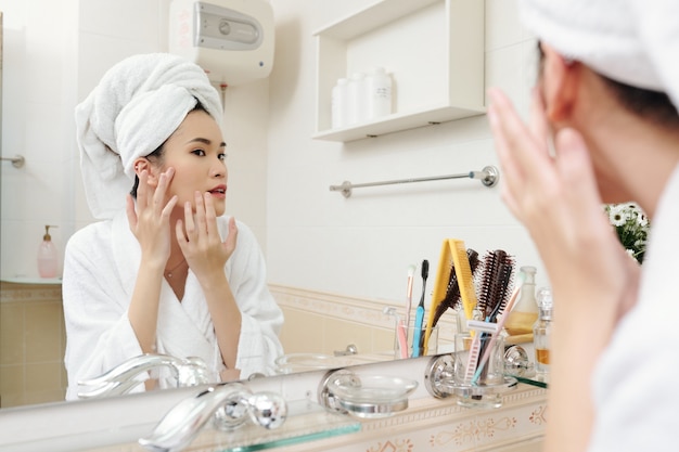 아침 샤워를 한 후 욕실 거울에 서서 피부의 흠집을 바라보는 젊은 아시아 여성