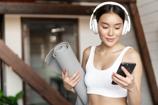 Young asian woman standing at home in headphones listening music and going workout on floor mat prac...