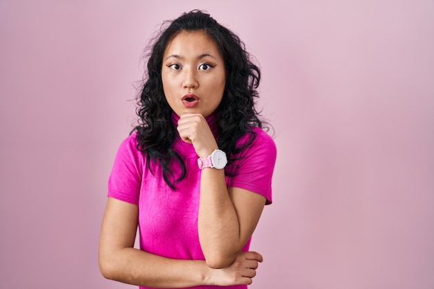 Young asian woman standing over pink background looking fascinated with disbelief, surprise and amazed expression with hands on chin