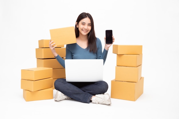 Young asian woman startup small business freelance holding parcel box, mobile phone and computer laptop and sitting on floor isolated, Online marketing packing box delivery concept