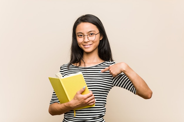 Young asian woman student holding a book person pointing by hand to a shirt blank space, proud and confident