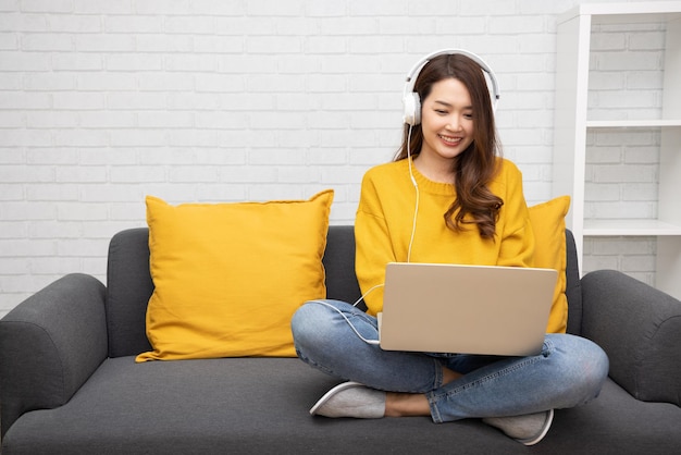 Young Asian woman student with wireless headset sitting with laptop on sofa