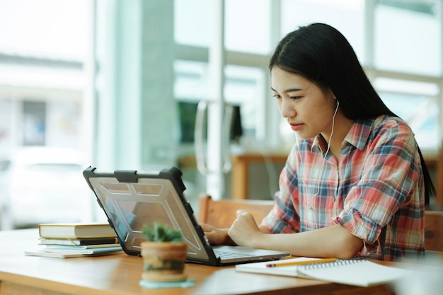 젊은 아시아 여성은 노트북 컴퓨터 앞에서 공부하고 offsitexA에서 헤드폰을 사용합니다.