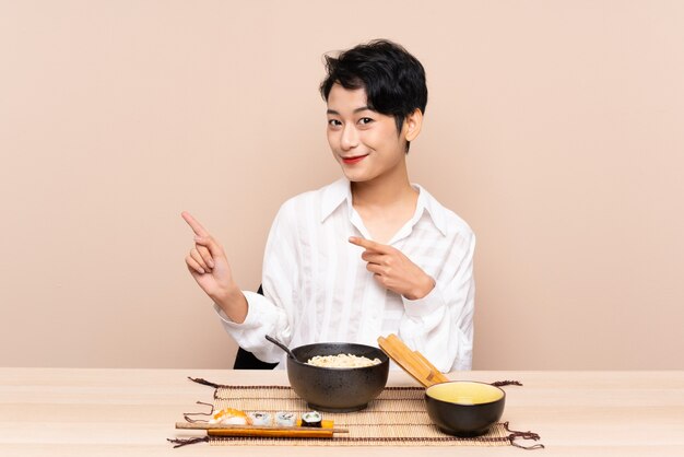 Young Asian woman in a table with bowl of noodles and sushi pointing finger to the side