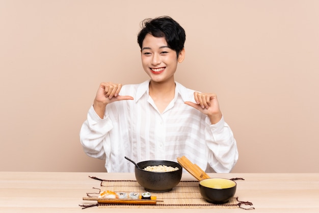 Young Asian woman in a table with bowl of noodles and sushi proud and self-satisfied