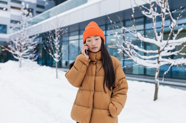 눈 덮인 겨울날 캠퍼스에서 걸어가는 전화 통화를 하는 젊은 아시아 여성