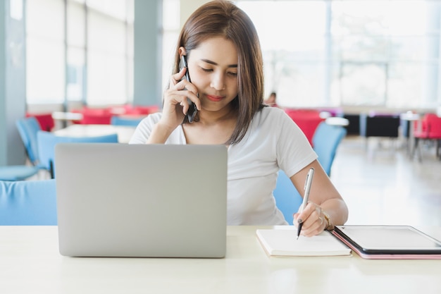 Foto giovane donna asiatica parlando con qualcuno sul suo telefono cellulare e note sul notebook.