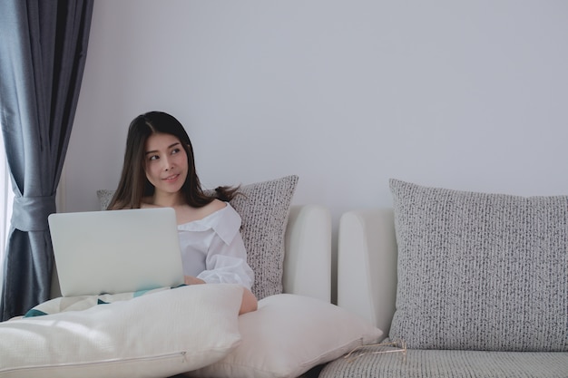 young asian woman thinking and using laptop on sofa bed in living room.