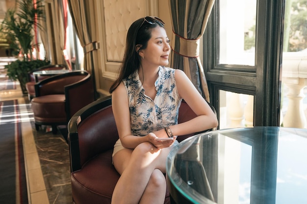 Young asian woman traveler waiting someone while sitting at lobby area of modern hotel. patiently girl tourist enjoy garden view and looking through window. smiling lady relax indoors on comfort sofa