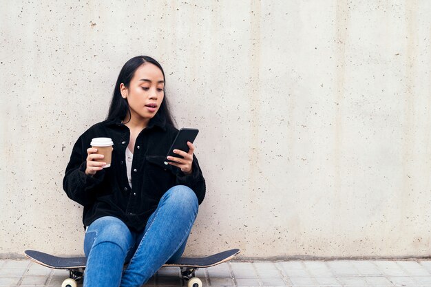 콘크리트 벽에 기대어 스케이트보드를 타고 야외에 앉아 전화를 하는 젊은 아시아 여성