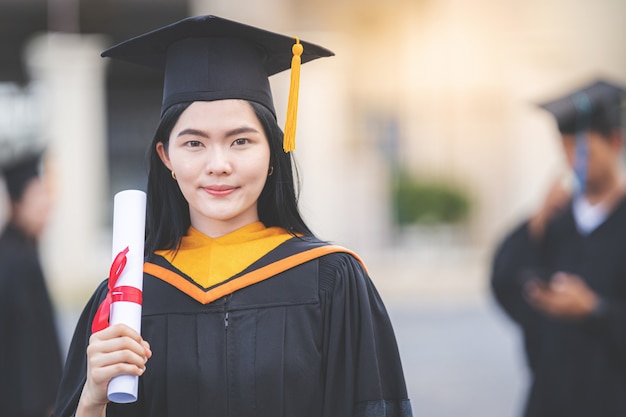 Una giovane donna asiatica laureata in abito da laurea e sparviere in possesso di un certificato di laurea si trova di fronte all'edificio universitario dopo aver partecipato all'inizio del college