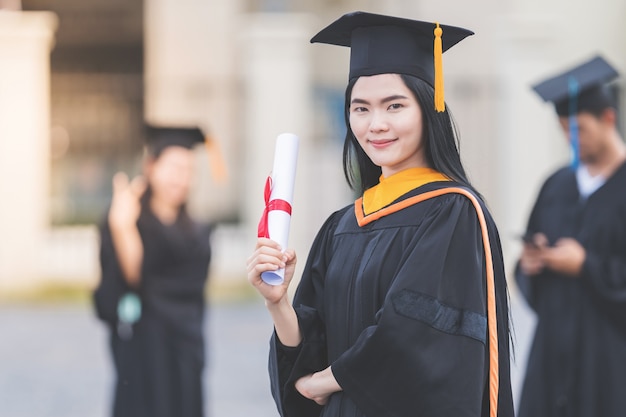 A young asian woman university graduate in graduation gown and
mortarboard holds a degree certificate stands in front of the
university building after participating in college
commencement