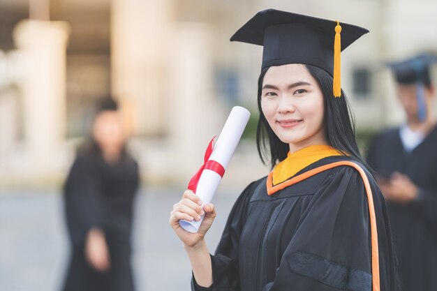 Una giovane donna asiatica laureata in abito da laurea e sparviere in possesso di un certificato di laurea si trova di fronte all'edificio universitario dopo aver partecipato all'inizio del college