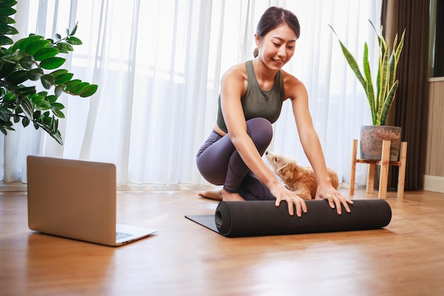 Young asian woman unrolling roll black yoga mat with her dog at\
home new normal lifestyle concept