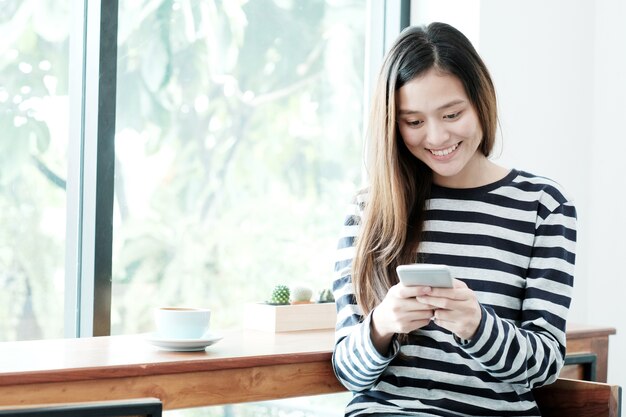 窓のカフェの背景で座っている間にスマートフォンを使用している若いアジアの女性