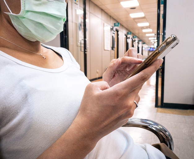 스마트폰을 사용하고 병원에서 수술용 마스크를 쓰고 있는 젊은 아시아 여성. 코로나바이러스, Covid19 전염병의 의료. 전화에 텍스트를 작성 하는 소녀. 보호 마스크는 바이러스 위험을 줄입니다