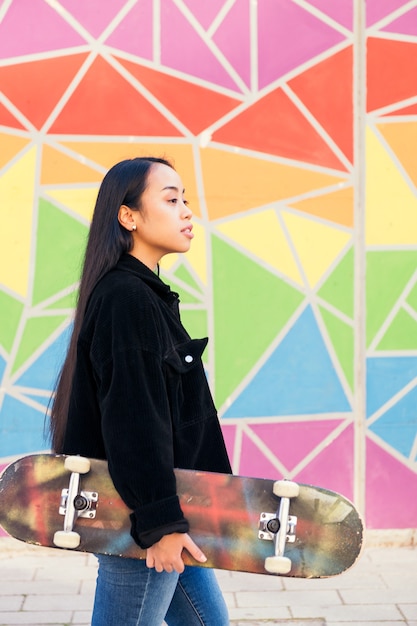 Молодая азиатская женщина гуляет со скейтбордом в руке перед красочной стеной