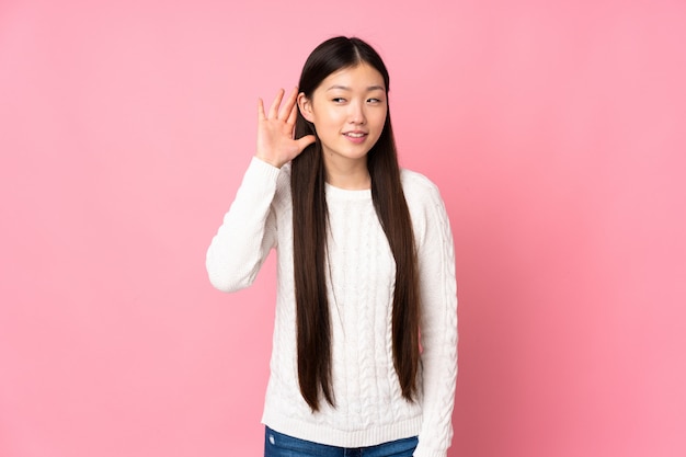 Young asian woman over wall listening to something by putting hand on the ear