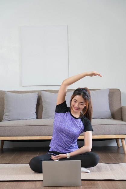 Молодая азиатская женщина смотрит фитнес-видео на ноутбуке и протягивает руку, чтобы расслабиться после тренировки