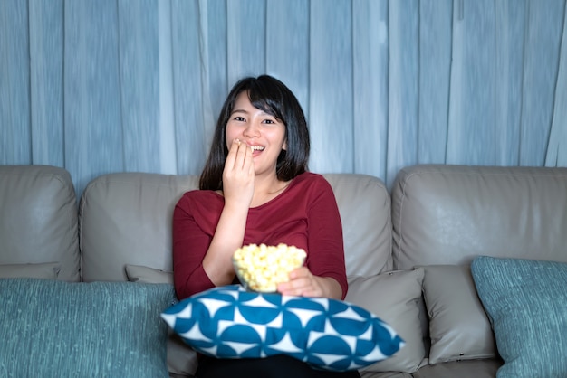 Young asian woman watching television suspense movie or news looking happy eating popcorn late night at home living room couch.