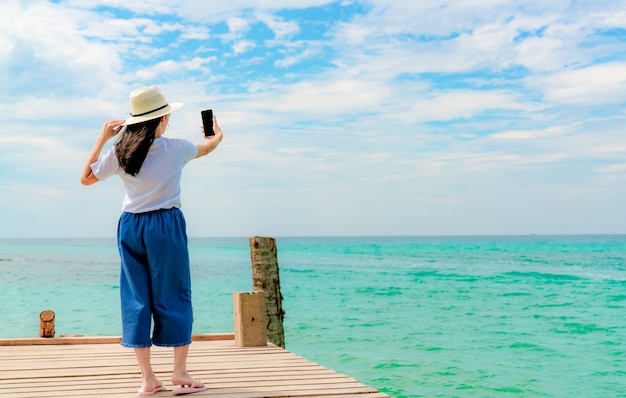 Young Asian woman wear straw hat in casual style use smartphone taking selfie at wooden pier. Summer vacation at tropical paradise beach. Happy girl travel on holiday.