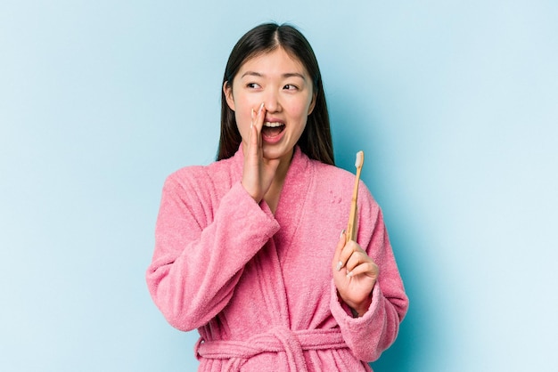 Young asian woman wearing a bathrobe brushing teeth isolated on pink background shouting and holding palm near opened mouth