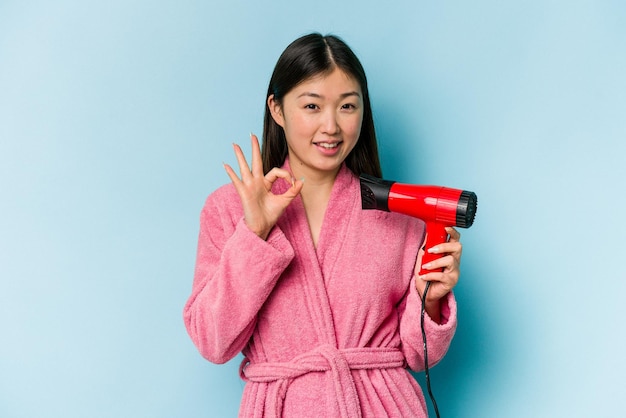 バスローブを着て、ピンクの背景に分離されたヘアドライヤーを保持している若いアジアの女性は、陽気で自信を持ってOKジェスチャーを示しています