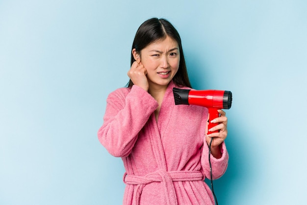 バスローブを着て、手で耳を覆うピンクの背景に分離されたヘアドライヤーを保持している若いアジアの女性