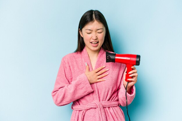 バスローブを着て、ピンクの背景に分離されたヘアドライヤーを保持している若いアジアの女性は、胸に手を置いて大声で笑う