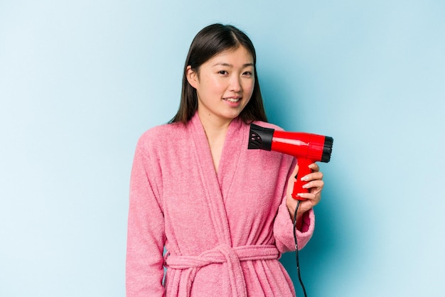 Young asian woman wearing a bathrobe and holding hairdryer isolated on pink background looks aside smiling cheerful and pleasant