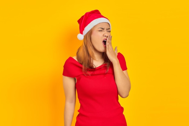 Young Asian woman wearing Christmas hat looking sleepy and covering mouth with hands