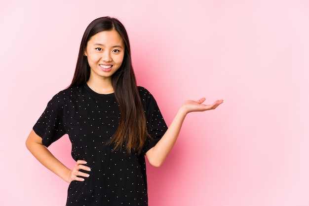 Giovane donna asiatica che indossa vestiti eleganti isolati mostrando uno spazio di copia su una palma e tenendo un'altra mano sulla vita.