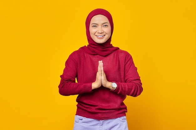 Young asian woman wearing headscarf gives greeting hands at with big smile on her face isolated on yellow background