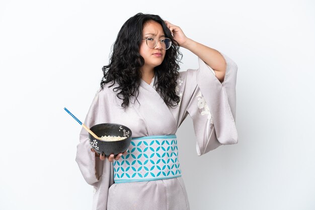 Молодая азиатская женщина в кимоно изолирована на белом фоне с сомнениями и смущенным выражением лица, держа миску лапши с палочками для еды