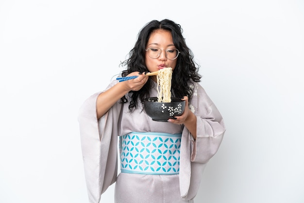 Giovane donna asiatica che indossa il kimono isolato su sfondo bianco tenendo una ciotola di tagliatelle con bacchette di sabbia che soffia perché sono calde