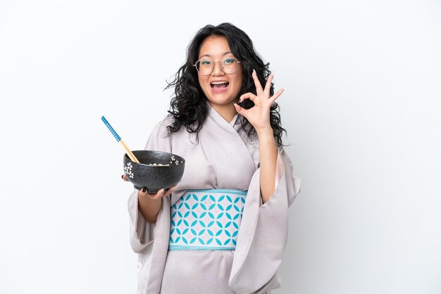 Young asian woman wearing kimono isolated on white background showing ok sign with fingers while holding a bowl of noodles with chopsticks