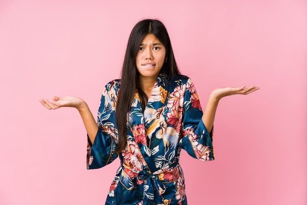 Young asian woman wearing a kimono pajamas confused and doubtful shrugging shoulders to hold a copy space.