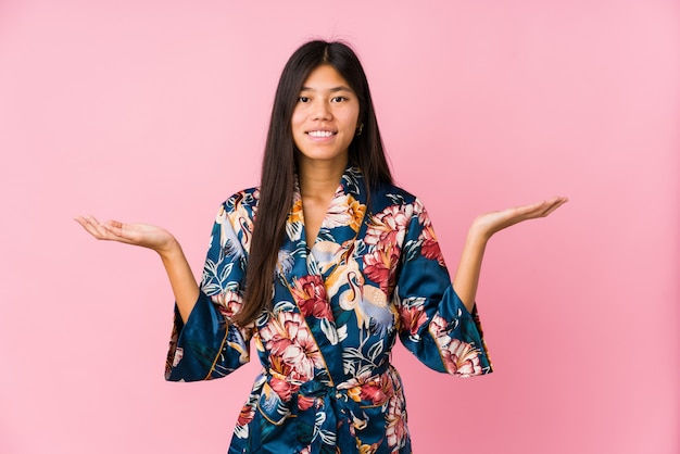 Young asian woman wearing a kimono pajamas makes scale with arms, feels happy and confident.